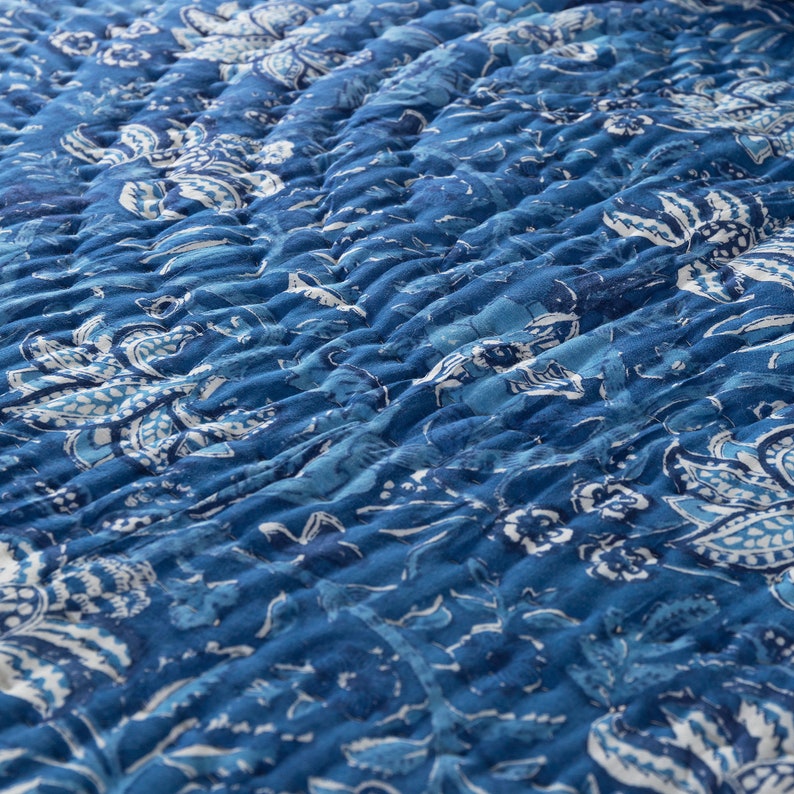 May include: Close-up of a quilted textile with a floral pattern in shades of blue and white. The fabric has a textured, rippled appearance, with visible stitching creating a raised effect. The design features stylized flowers and leaves.