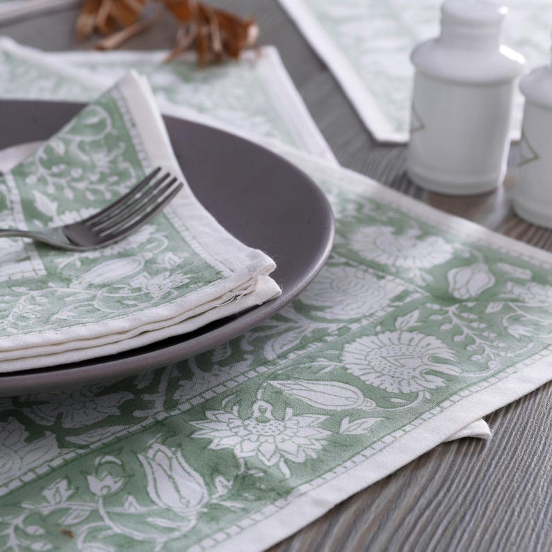 Pode incluir: Um tapete de mesa verde e branco com padr&atilde;o floral e uma toalha de mesa verde e branca dobrada em um prato cinza. O tapete de mesa &eacute; feito de algod&atilde;o e tem uma borda branca.