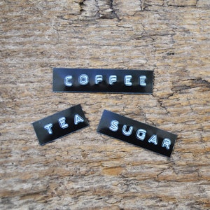 May include: Three black rectangular labels with white lettering on a wooden surface. The labels read "COFFEE", "TEA", and "SUGAR".