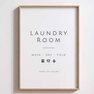 Op de afbeelding: Een wit ingelijst bord met de tekst "Laundry Room" in zwarte letters. Onder de tekst staat "Wash - Dry - Fold" met was symbolen voor wassen, drogen en ophangen van kleding. Het bord zegt ook "Open 24 Hours".