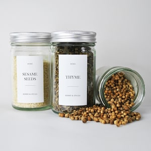 Puede incluir: Tres frascos de vidrio con tapas plateadas, etiquetados como "Sesame Seeds" y "Thyme", y "Herbs & Spices". Un frasco está volcado, derramando semillas marrones. Los frascos contienen semillas de sésamo, tomillo y semillas de cilantro.