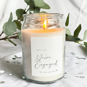 Könnte beinhalten: Ein klares Glas-Kerzengefäß mit einem weißen Etikett, auf dem "Smells like You're Engaged" steht, darunter der Text "Mr & Mrs Page to be". Die Kerze ist angezündet und die Flamme ist sichtbar.