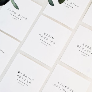 May include: White square labels with black text for laundry products. Labels include "Hand Soap", "Washing Pods", "Stain Remover", "Scent Boosters", "Washing Powder", and "Laundry Detergent". Green leaves are in the background.