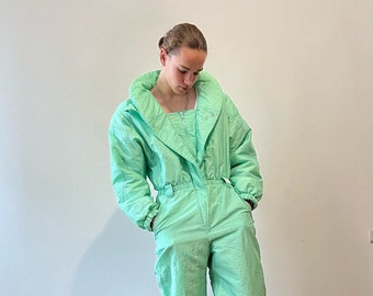 May include: A light green one-piece jumpsuit with a high collar and a zipper closure. The jumpsuit has a drawstring waist and is made of a lightweight fabric. The model is wearing white sneakers.