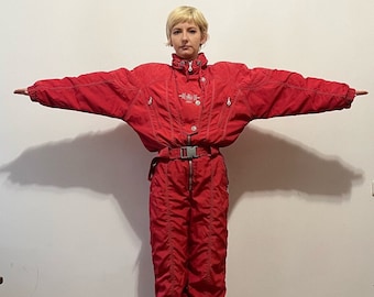May include: A red ski suit with a white zipper and a white belt. The suit has a logo on the chest that says "Alpina".