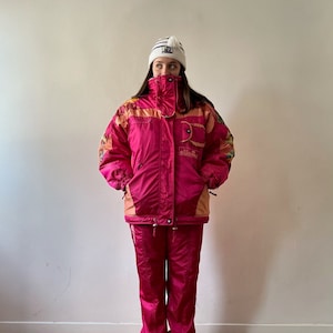 May include: A pink ski suit with a white and orange patterned design. The jacket has a hood and a zipper closure. The pants have an elastic waistband. The suit is made of a shiny, nylon fabric.