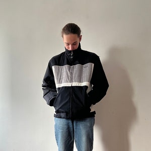 May include: Black and gray track jacket with a white panel across the chest. The jacket has a high collar and a zipper closure. The sleeves are black, and the jacket is paired with blue jeans.