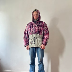 May include: A hooded jacket with a unique pattern in shades of pink, purple, and white, paired with a light green lower section. The jacket is worn with blue jeans and black and white sneakers. The jacket has a drawstring at the waist.