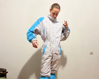 May include: A white ski jumpsuit with blue accents. The jumpsuit has a zipper down the front and a high collar. The model is wearing the jumpsuit with a pair of brown sneakers.