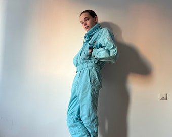 May include: A light blue ski jumpsuit with a high collar and a zipper closure. The jumpsuit has a belt and is made of a nylon fabric. The person is wearing black sneakers.