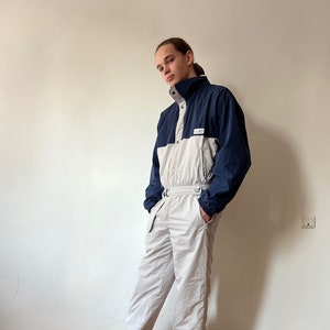 May include: A white and blue ski jumpsuit with a zipper closure and a high collar. The jumpsuit has a patch on the chest with the word "Salomon" printed on it. The person is wearing patterned sneakers.