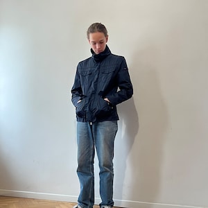 May include: A navy blue jacket with a high collar and multiple pockets. The jacket is worn over a white shirt and blue jeans. The person is wearing black sneakers.