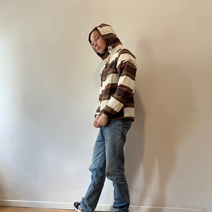 May include: A person wearing a brown and white striped hooded jacket with blue jeans and purple and white sneakers.