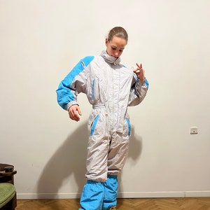 May include: A white ski jumpsuit with blue accents. The jumpsuit has a zipper down the front and a high collar. The model is wearing the jumpsuit with a pair of brown sneakers.