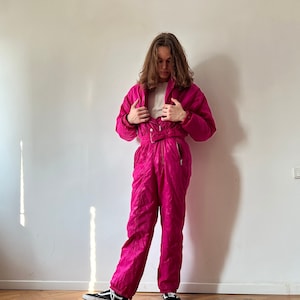 May include: A pink ski suit with a zipper and a belt. The suit is made of a shiny, quilted fabric. The person is wearing black sneakers.