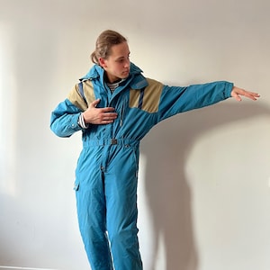 May include: A blue ski jumpsuit with brown accents and a hood. The jumpsuit has a zipper closure and pockets. The person is wearing the jumpsuit with a striped shirt underneath.