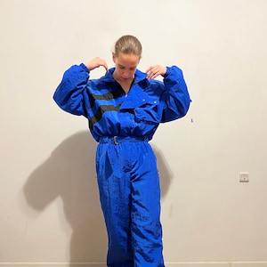May include: A blue nylon jumpsuit with black stripes and a belt. The jumpsuit has a zipper closure and a pocket on the chest. The person is wearing tan sneakers.