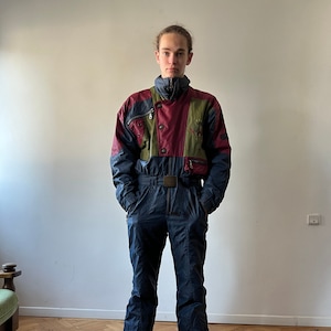 May include: A blue, green, and red ski suit with a zipper and a belt. The suit has multiple pockets and a high collar.