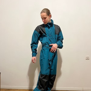 May include: Teal and black vintage jumpsuit with a high collar and button closure. The jumpsuit features black panels on the shoulders and lower legs. The model is wearing tan sneakers. A retro-style one-piece garment.