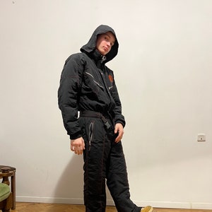May include: A black ski suit with red stitching and a hood. The suit has a zipper closure and pockets. The person is wearing brown shoes.