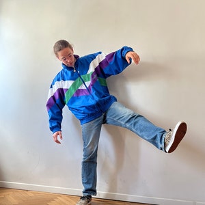 Può includere: Una persona indossa una giacca a vento blu, viola, bianca e verde con una cerniera e una striscia bianca sul davanti. Indossa jeans blu e scarpe da ginnastica marroni e nere.