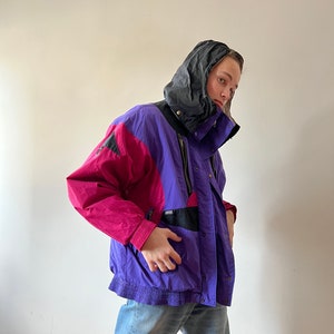 Może przedstawiać: Fioletowa, różowa i czarna kurtka narciarska z czarnym kapturem. Kurtka zapinana na zamek błyskawiczny i posiada wiele kieszeni.