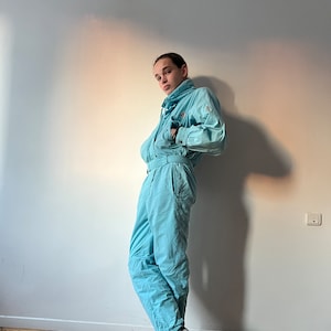 May include: A light blue ski jumpsuit with a high collar and a zipper closure. The jumpsuit has a belt and is made of a nylon fabric. The person is wearing black sneakers.