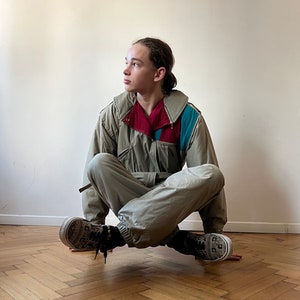 May include: A person wearing a beige jumpsuit with a red, blue, and green color block design on the jacket. The person is sitting on a wooden floor with their legs crossed.