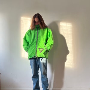 May include: A bright green jacket with a zipper closure and a logo on the left chest. The jacket is worn over blue jeans and black sneakers.