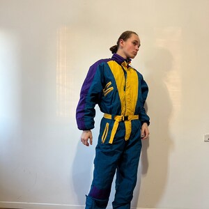 May include: A blue, yellow, and purple ski suit with a yellow belt. The suit has a zipper up the front and a hood.