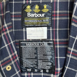 Peut inclure: Une &eacute;tiquette d'entretien Barbour International Suit avec des instructions pour laver, s&eacute;cher et ranger le v&ecirc;tement. L'&eacute;tiquette est fix&eacute;e &agrave; une veste en tissu &eacute;cossais bleu marine et blanc. L'&eacute;tiquette comprend le texte "Barbour Care" et "100% Coton/Coton Minimum".