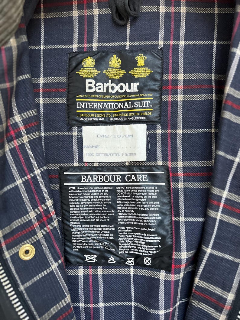 Peut inclure: Une &eacute;tiquette d'entretien Barbour International Suit avec des instructions pour laver, s&eacute;cher et ranger le v&ecirc;tement. L'&eacute;tiquette est fix&eacute;e &agrave; une veste en tissu &eacute;cossais bleu marine et blanc. L'&eacute;tiquette comprend le texte "Barbour Care" et "100% Coton/Coton Minimum".