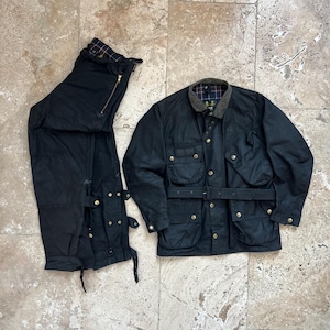 Peut inclure: Une veste en coton cir&eacute; noir avec une ceinture et plusieurs poches. La veste est pos&eacute;e &agrave; plat sur une surface carrel&eacute;e. &Agrave; c&ocirc;t&eacute; de la veste se trouve un pantalon en coton cir&eacute; noir avec une fermeture &eacute;clair et plusieurs boucles.