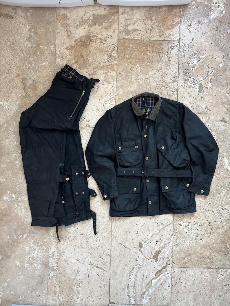 Peut inclure: Une veste en coton cir&eacute; noir avec une ceinture et plusieurs poches. La veste est pos&eacute;e &agrave; plat sur une surface carrel&eacute;e. &Agrave; c&ocirc;t&eacute; de la veste se trouve un pantalon en coton cir&eacute; noir avec une fermeture &eacute;clair et plusieurs boucles.