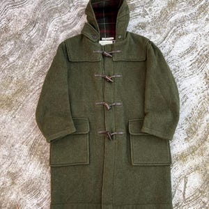 May include: An olive green duffel coat with a hood and toggle fastenings. The coat has two front pockets and a plaid lining visible in the hood. The coat is made of a thick, wool-like material.