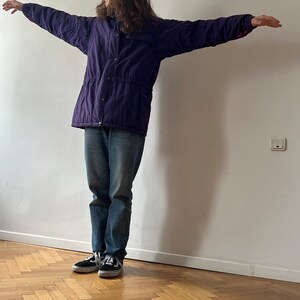 Pu&ograve; includere: Una persona indossa una giacca viola con fodera rossa, jeans blu e scarpe da ginnastica nere. La persona &egrave; in piedi con le braccia distese davanti a un muro bianco.
