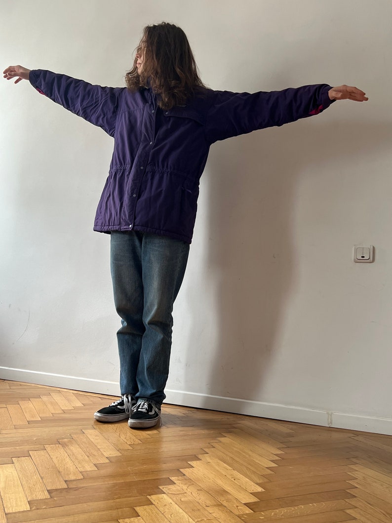 Pu&ograve; includere: Una persona indossa una giacca viola con fodera rossa, jeans blu e scarpe da ginnastica nere. La persona &egrave; in piedi con le braccia distese davanti a un muro bianco.