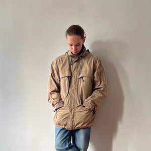 May include: A tan jacket with a zipper and multiple pockets. The jacket has a hood and is made of a water-resistant material.