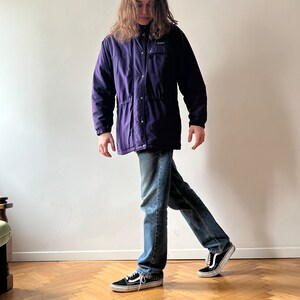 Pu&ograve; includere: Una persona indossa una giacca viola con cerniera e chiusura a pressione, jeans blu e scarpe da ginnastica nere. La giacca ha un logo sul petto sinistro.