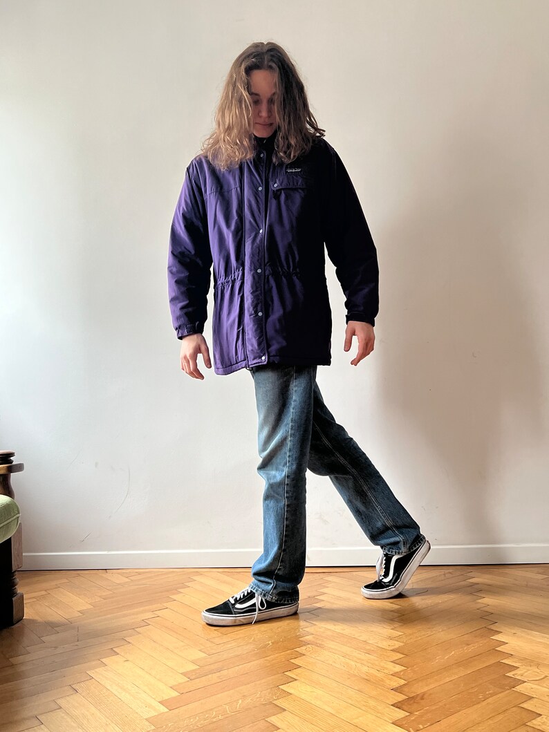Pu&ograve; includere: Una persona indossa una giacca viola con cerniera e chiusura a pressione, jeans blu e scarpe da ginnastica nere. La giacca ha un logo sul petto sinistro.