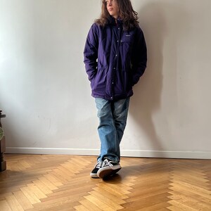Pu&ograve; includere: Una giacca viola con cappuccio, chiusura a pressione e cerniera. La giacca ha una toppa sul petto sinistro con la scritta "Patagonia". La persona indossa jeans blu e scarpe da ginnastica nere.