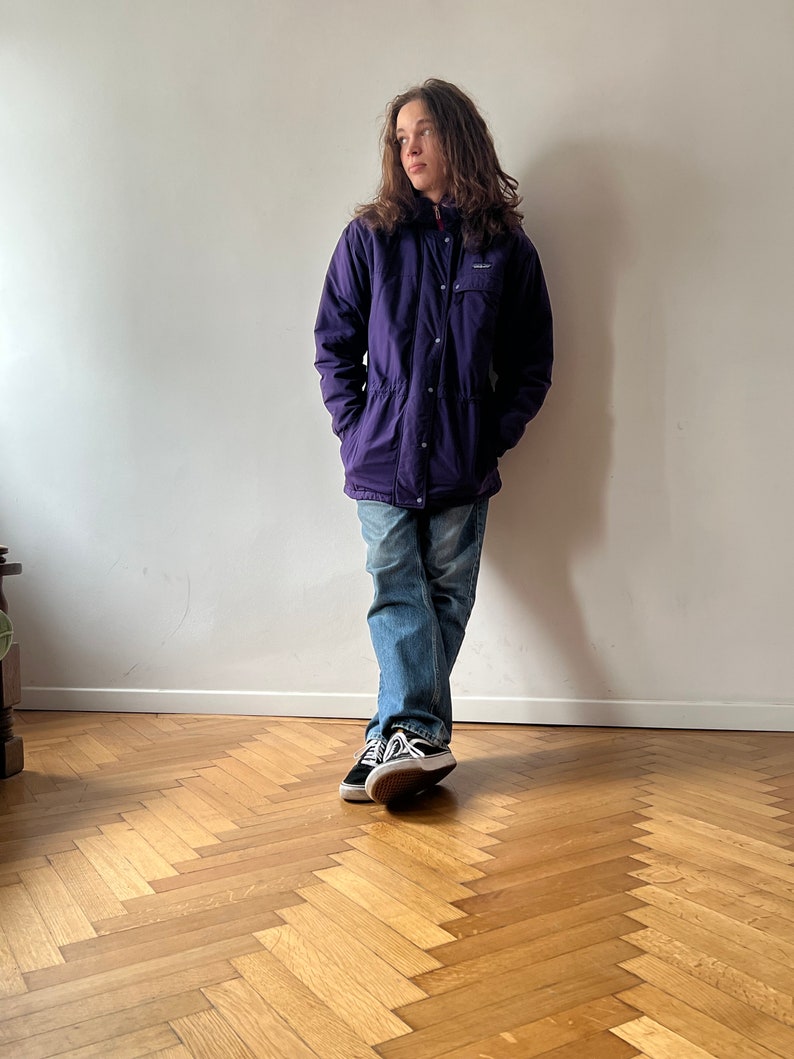 Pu&ograve; includere: Una giacca viola con cappuccio, chiusura a pressione e cerniera. La giacca ha una toppa sul petto sinistro con la scritta "Patagonia". La persona indossa jeans blu e scarpe da ginnastica nere.