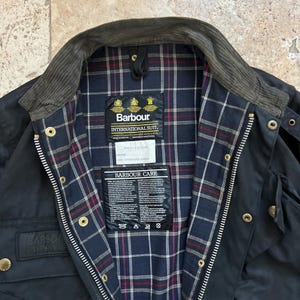 Peut inclure: Une veste Barbour International Suit bleu marine avec un col en velours c&ocirc;tel&eacute; et une doublure &agrave; carreaux. La veste a une fermeture &eacute;clair et plusieurs poches. L'&eacute;tiquette Barbour Care est visible &agrave; l'int&eacute;rieur de la veste.