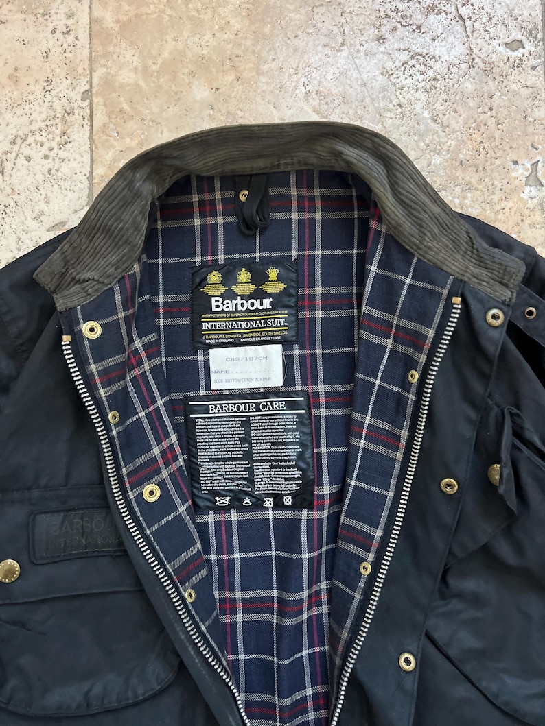 Peut inclure: Une veste Barbour International Suit bleu marine avec un col en velours c&ocirc;tel&eacute; et une doublure &agrave; carreaux. La veste a une fermeture &eacute;clair et plusieurs poches. L'&eacute;tiquette Barbour Care est visible &agrave; l'int&eacute;rieur de la veste.