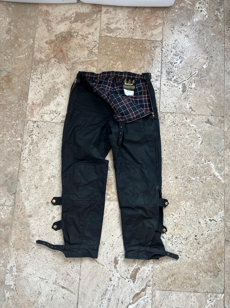 Peut inclure: Pantalon en coton cir&eacute; noir Barbour avec des bretelles r&eacute;glables et une doublure en tartan. Le pantalon a deux grandes poches sur le devant et deux petites poches &agrave; l'arri&egrave;re.