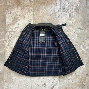 Peut inclure: Une veste Barbour vert fonc&eacute; avec une doublure en tartan noir et rouge. La veste a une fermeture &eacute;clair et un col boutonn&eacute;. Le logo Barbour est visible &agrave; l'int&eacute;rieur de la veste.