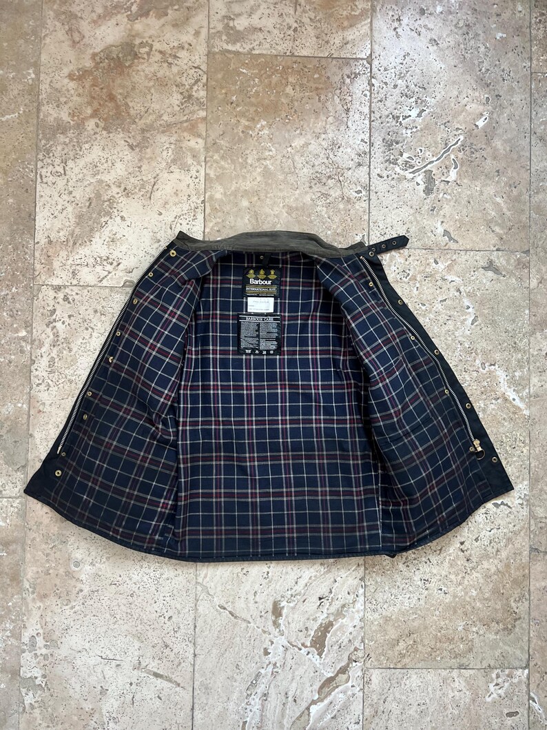 Peut inclure: Une veste Barbour vert fonc&eacute; avec une doublure en tartan noir et rouge. La veste a une fermeture &eacute;clair et un col boutonn&eacute;. Le logo Barbour est visible &agrave; l'int&eacute;rieur de la veste.