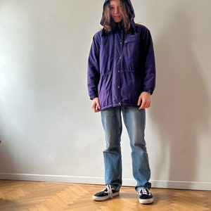 Pu&ograve; includere: Una giacca viola con cappuccio, cerniera e chiusura a bottoni. La giacca ha una vita con coulisse e due tasche sul davanti.
