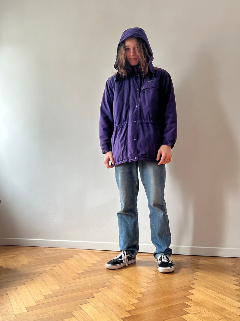 Pu&ograve; includere: Una giacca viola con cappuccio, cerniera e chiusura a bottoni. La giacca ha una vita con coulisse e due tasche sul davanti.