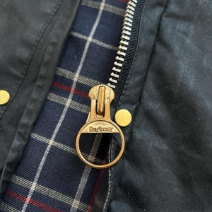 Peut inclure: Gros plan sur une fermeture &eacute;clair de veste Barbour noire avec un tirette en laiton sur laquelle est inscrit "Barbour". La fermeture &eacute;clair est partiellement ouverte, r&eacute;v&eacute;lant une doublure en tissu &agrave; carreaux bleu et blanc.